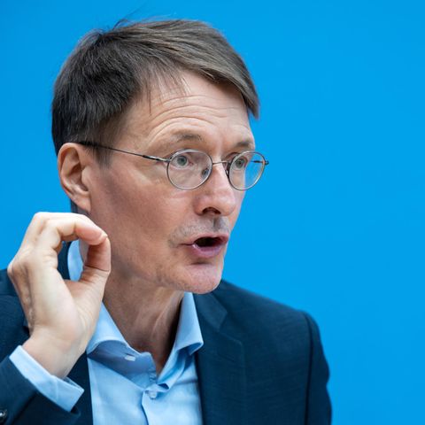 Ein weißer, älterer Mann mit Metallbrille und angegrautem Seitenscheitel sitzt im Anzug vor einer blauen Wand und gestikuliert