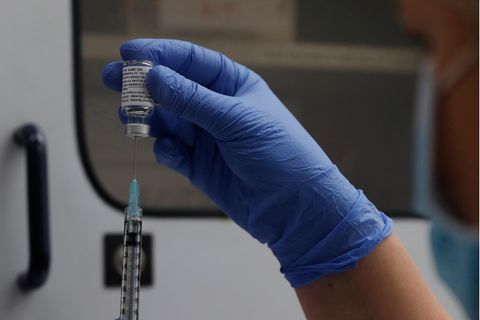 Eine Hand in blauem Gumihandschuh hält eine Ampulle mit Novavx über Kopf, während von unten eine Spritze hineinsticht