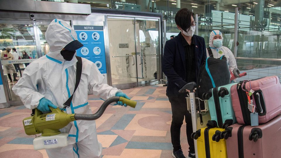 Eine Person in weißem Schutzanzug sprüht Gepäck und Ankömmlinge am Flughafen ein