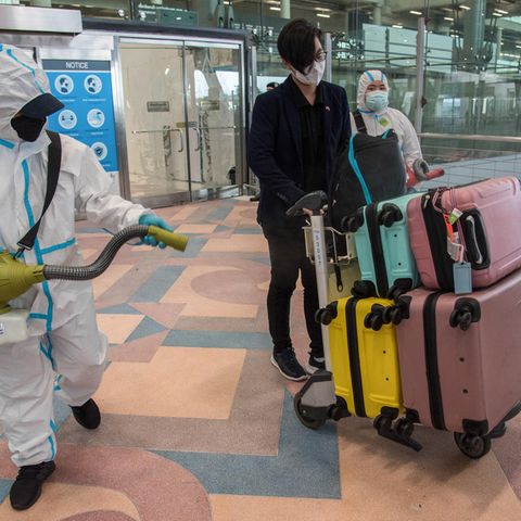 Eine Person in weißem Schutzanzug sprüht Gepäck und Ankömmlinge am Flughafen ein