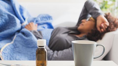 Frau liegt krank auf dem Sofa. Im Vordergrund: eine Tasse, Hustensaft und ein Fieberthermometer