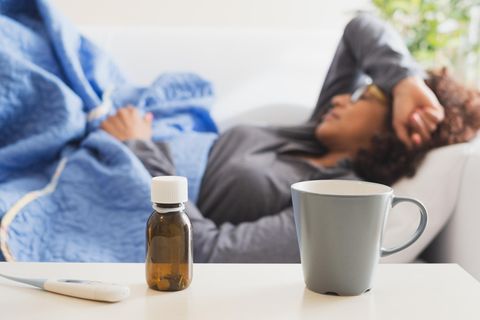 Frau liegt krank auf dem Sofa. Im Vordergrund: eine Tasse, Hustensaft und ein Fieberthermometer