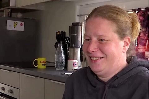 Eine blonde, weiße Frau im grauen Kapuzenpullover lächelt, während sie in einem Tiny House sitzt