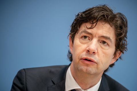Christian Drosten sitzt im Anzug vor einer blauen Wand