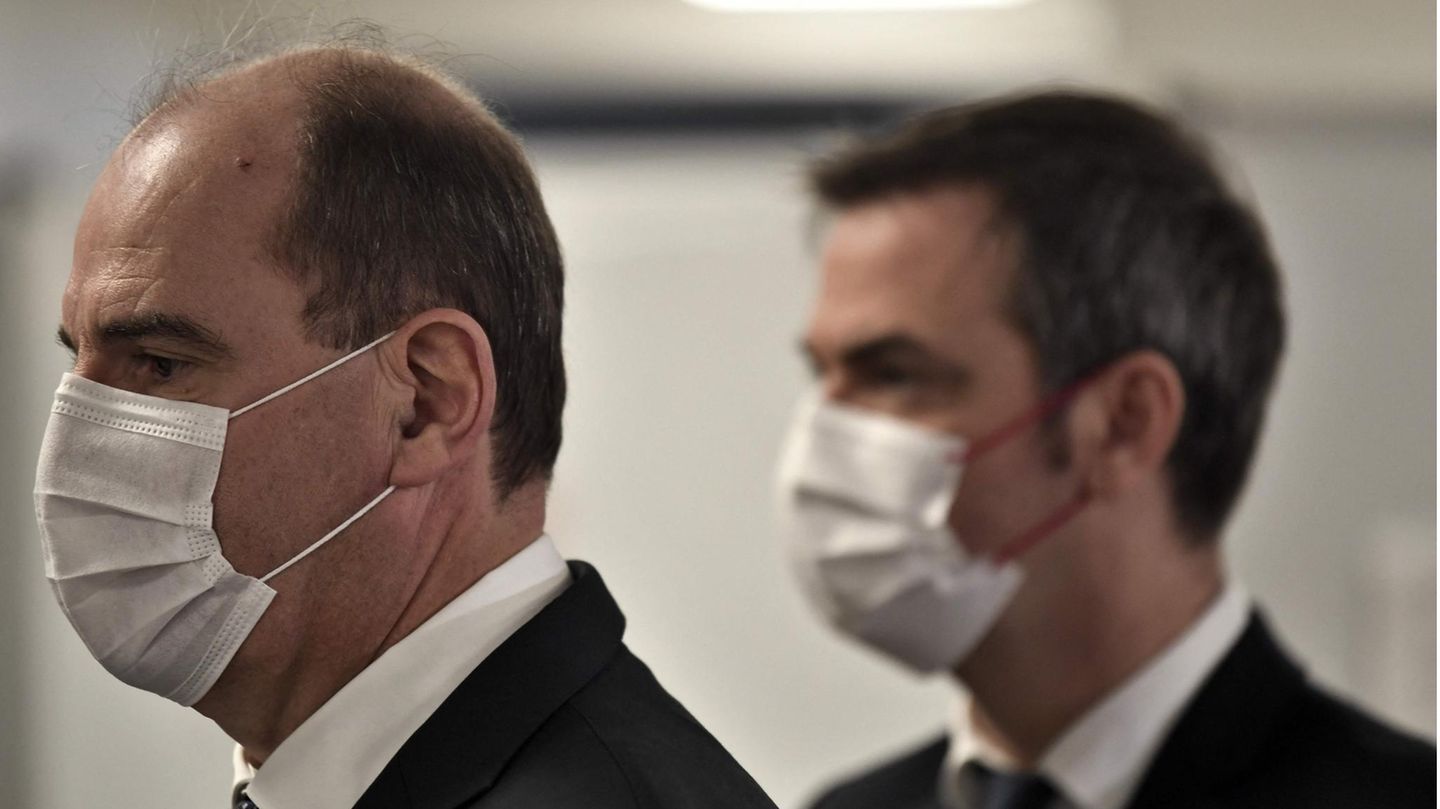 Zwei weiße ältere Männer in schwarzen Anzügen und mit weißen OP-Masken stehen auf dem Flur einer medizinischen Einrichtung