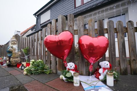 Vor einem grau-braunen Lattenzaun stehen weiße Grablichter neben zwei weißen Weihnachtsteddys, die rote Herzballons halten