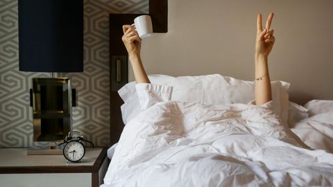 Eine Frau streckt die Hand, mit der sie eine Kaffeetasse hält, aus der Bettdecke hervor