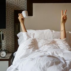 Eine Frau streckt die Hand, mit der sie eine Kaffeetasse hält, aus der Bettdecke hervor