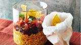 Burrito-Schicht-Salat      Zutaten:  250 g Naturreis (ersatzweise »Dinkel wie Reis«), 1 Bio-Limette (ersatzweise BioZitrone), 500 g Tomaten, 1 Bund Schnittlauch, 3 EL Ahornsirup, Salz, Pfeffer, 1 Zwiebel, 1 Dose schwarze Bohnen, (240 g Abtropfgewicht, ersatzweise Kidneybohnen), 1 EL neutrales Pflanzenöl, 1 EL Tomatenmark, ¼ TL gemahlener Kreuzkümmel, ¼ TL gemahlener Koriander, 2 Msp. Cayennepfeffer, ½ TL gemahlene Kurkuma, 2 EL Weißwein- oder Apfelessig, 2 TL geröstetes Sesamöl, ½ Bund Koriander, 3 Handvoll Tortillachips      Zubereitung:  1. Den Reis in ein feines Sieb geben und mit kaltem Wasser abwa - schen, bis das Wasser klar bleibt. Dann in der doppelten Menge Wasser nach Packungsanweisung garen.  2. Für die Salsa die Limette heiß abwaschen, trocken tupfen, die Schale abreiben und den Saft auspressen. Die Tomaten waschen, vom Stielansatz befreien und entkernen. Das Fruchtfleisch fein würfeln. Den Schnittlauch waschen, trocken schütteln und in feine Röll - chen schneiden. Schnittlauch, 1 EL Ahornsirup und Limettensaft zu den Tomatenwürfeln geben und unterheben, die Salsa mit Salz und Pfeffer abschmecken.  3. Die Zwiebel schälen und würfeln. Die Bohnen durch ein Sieb abgie - ßen und abtropfen lassen. Das Öl in einer Pfanne erhitzen und die Zwiebeln darin goldbraun anbraten. Bohnen und Tomatenmark da - zugeben und unter Rühren 1 Min. anrösten. Kreuzkümmel, Korian - der und Cayennepfeffer dazugeben, umrühren und mit 100 ml Was - ser ablöschen. 30 Sek. köcheln lassen, bis die Sauce eindickt, dann vom Herd ziehen und mit Salz abschmecken.  4. Den Reis mit Kurkuma, Essig, Sesamöl und restlichem Ahornsirup (2 EL) mischen, mit etwas Salz abschmecken und zum Mitnehmen auf 4 Schraubgläser oder Boxen verteilen. Die Bohnen und Tomaten obenauf schichten. Koriander waschen, trocken schütteln und über die Bohnen streuen. Verschließen und kalt stellen. Die Chips ge - trennt verpacken und zum Salat servieren.