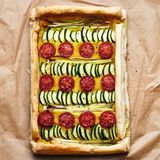 Blätterteigtarte mit Fava und Gemüse      Zutaten:  200 g gelbe Schälerbsen (Fava, ersatzweise gelbe Linsen), 2 Lorbeerblätter, 1 Zwiebel, 2 Knoblauchzehen, 1 kleine Bio-Zitrone, 50–80 ml ungesüßter Pflanzendrink, 4 EL natives Olivenöl, ½ TL gemahlene Kurkuma, Salz, ½ Zucchino, 2 Frühlingszwiebeln, 6 Kirschtomaten, 1 Packung veganer Blätterteig (275 g), schwarzer Pfeffer aus der Mühle      Zubereitung:  1. Die Erbsen 3 Std. in kaltem Wasser einweichen. Das Einweichwasser danach abgießen und die Erbsen mit den Lorbeerblättern in der doppelten Menge Wasser aufkochen. Ohne Deckel in ca. 20 Min. weich kochen (Achtung, es schäumt!).  2. Währenddessen die Zwiebel und den Knoblauch schälen und grob würfeln. Die Zitrone heiß abwaschen, trocknen, die Schale abreiben und 3 EL Saft auspressen. Die Erbsen abgießen, die Lorbeerblätter entfernen. Die weichen Erbsen mit Zwiebeln, Knoblauch, Zitronen - saft, 50 ml Pflanzendrink, Olivenöl, Kurkuma und etwas Salz fein cremig pürieren. Falls nötig, noch etwas mehr Pflanzendrink dazu - geben.  3. Zucchino, Frühlingszwiebeln und Tomaten waschen und abtropfen lassen. Vom Zucchino den Blüten- oder Stielansatz gerade abschnei - den und den Zucchino auf 10 cm Länge kürzen. Dann längs vier - teln, das Kerngehäuse herausschneiden und den Zucchino in 3 mm dicke Scheiben schneiden. Die Frühlingszwiebeln putzen und vom unteren, weißen Teil 10 cm lange Stücke abschneiden, diese vierteln. Die Tomaten halbieren.  4. Den Backofen auf 200° vorheizen. Ein Backblech mit Back - papier auslegen, den Blätterteig darauflegen und zu einer Fläche von 15 × 40 cm schneiden. Die kürzeren Seiten 1,5 cm, die langen Seiten 1 cm nach innen umschlagen und festdrücken. Vom rest - lichen Blätterteig 2 lange und 2 kurze Streifen abschneiden, sodass diese zu den Seitenlängen der Tarte passen.  5. Die Erbsencreme auf das große Blätterteigstück streichen, dabei die Ränder aussparen. Das Gemüse auf der Creme verteilen. Die Ränder der Tarte mit Wasser bestreichen, die übrigen Teigstreifen darauflegen und leicht andrücken. Die Tarte im Backofen (Mitte) 15 Min. backen, bis die Ränder der Tarte leicht gebräunt sind. Aus dem Ofen nehmen, mit etwas Salz und gemahlenem schwarzen Pfeffer aus der Mühle bestreuen. Dazu passt ein grüner Salat.