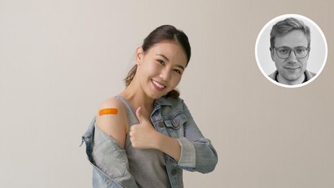 Eine asiatische aussehende Frau weist mit gehobenem Daumen auf das Pflaster auf ihrem Oberarm hin