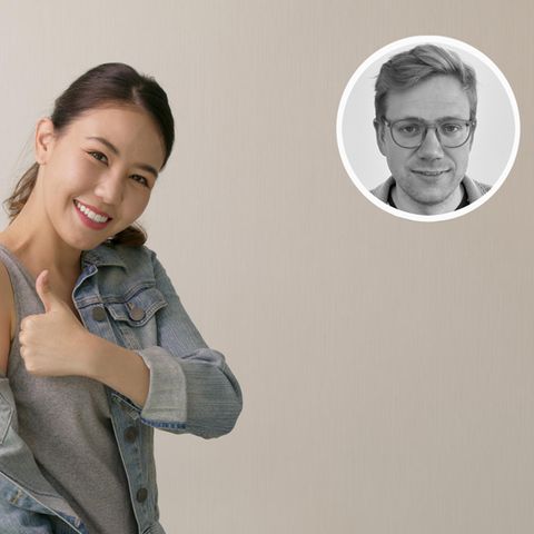 Eine asiatische aussehende Frau weist mit gehobenem Daumen auf das Pflaster auf ihrem Oberarm hin