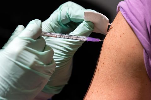 Zwei Hände in hellgrünen dünnen Gummihandschuhen stechen eine Spritze in den Oberarm eines weißen Mannes