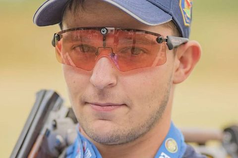 Ein junger Mann mit oranger Sortbrille und blauem Cap lächelt mit Gewehr über der Schulter in die Kamera