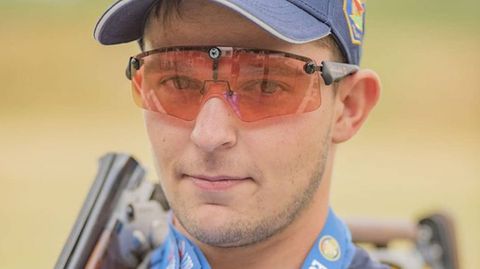 Ein junger Mann mit oranger Sortbrille und blauem Cap lächelt mit Gewehr über der Schulter in die Kamera