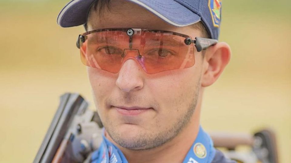 Ein junger Mann mit oranger Sortbrille und blauem Cap lächelt mit Gewehr über der Schulter in die Kamera