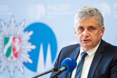 Ein weißer Mann mit grau-weißen lockigen Haaren sitzt in Anzug und Krawatte vor einer Wand mit dem Wappen der Polizei NRW