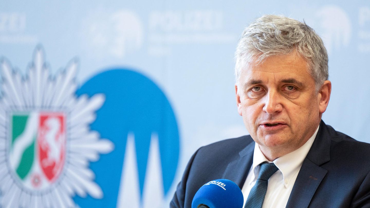 Ein weißer Mann mit grau-weißen lockigen Haaren sitzt in Anzug und Krawatte vor einer Wand mit dem Wappen der Polizei NRW