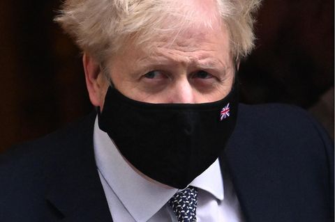 Ein weißer Mann mit strubbeligen weißblonden Haaren und schwarzer Maske mit Union Jack verlässt in Anzug und Krawatte ein Haus