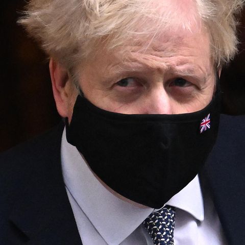 Ein weißer Mann mit strubbeligen weißblonden Haaren und schwarzer Maske mit Union Jack verlässt in Anzug und Krawatte ein Haus