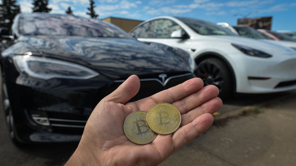 Eine Hand hält zwei Bitcoins vor Tesla-Fahrzeuge