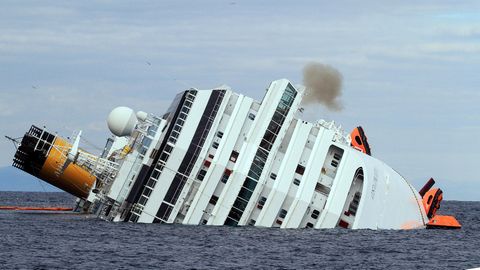 Ein weißes Kreuzfahrtschiff liegt mit Schlagseite im Meer. Auf der einen Seite hängen noch orangene Rettungsinseln