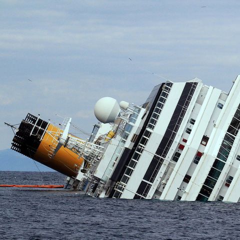 Ein weißes Kreuzfahrtschiff liegt mit Schlagseite im Meer. Auf der einen Seite hängen noch orangene Rettungsinseln