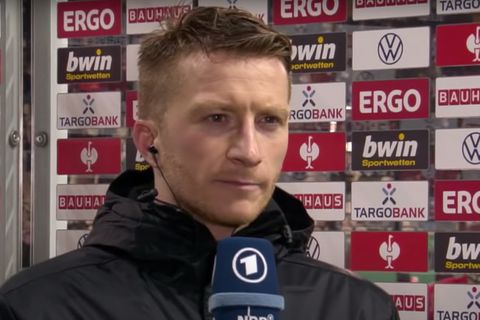 Vor einer Tafel mit Sponsorenlogos steht ein dunkelblonder, weißer Fußballer im schwarzen Parka und schaut ernst