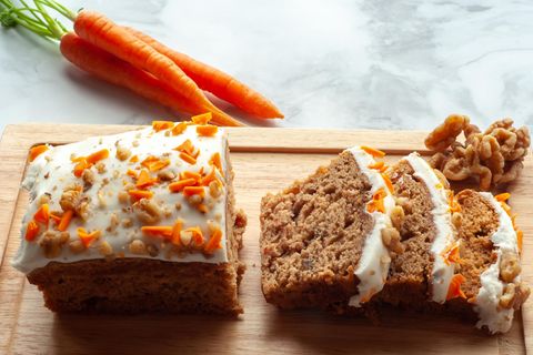 Ein angeschnittener Karottenkuchen auf einem Brett, dahinter ein Bund Möhren