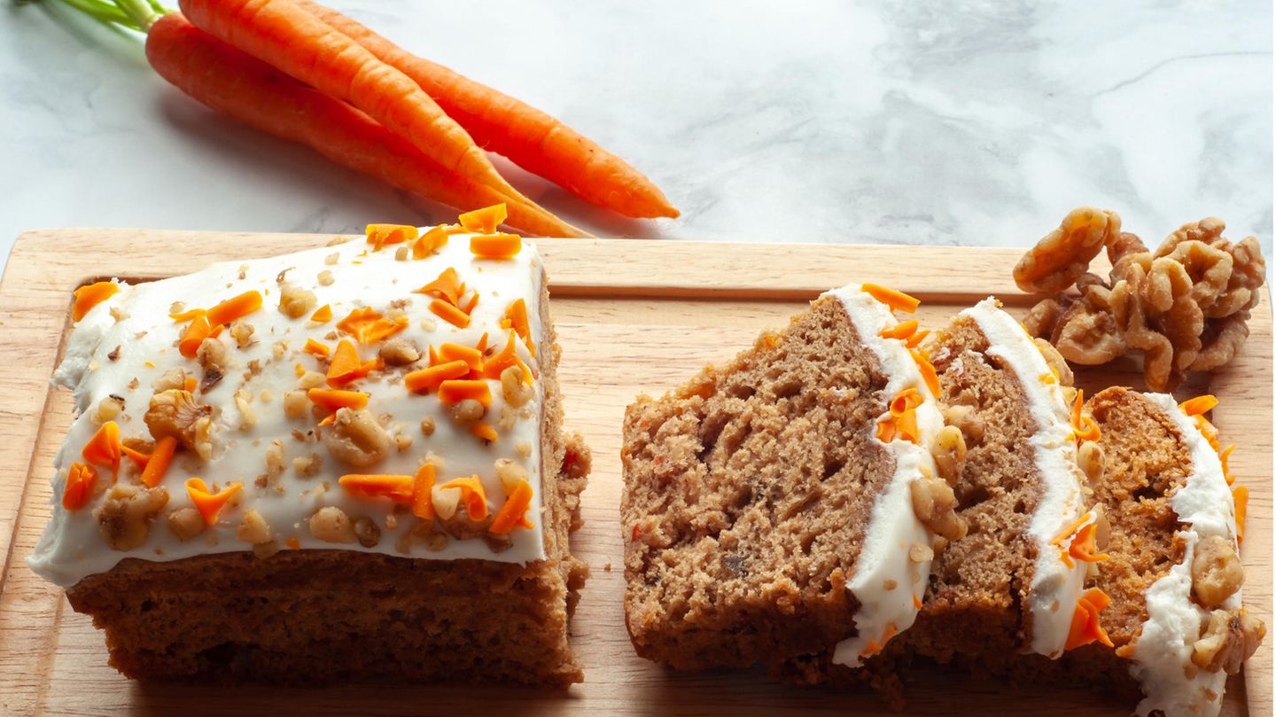 Ein angeschnittener Karottenkuchen auf einem Brett, dahinter ein Bund Möhren