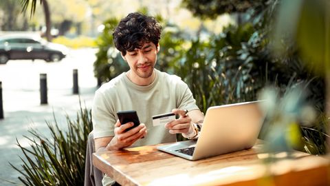 Ein junger Mann sitzt mit Handy und Kreditkarte am Laptop