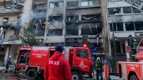 Rote Feuerwehrwagen mit weißen, griechischen Buchstaben darauf stehen vor einem rußgeschwärzten mehrstöckigem Wohnhaus
