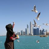 Eine Frau mit Kopftuch und in braun-rotem Gewand wirft Futter für Möwen in die Luft. Im Hintergrund das Meer und eine Skyline