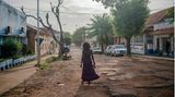 Eine schwarze Frau mit langem Rock geht durch eine Straße mit brüchigem Asphalt. Links und rechts stehen kleine, weiße Bungalows