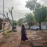 Eine schwarze Frau mit langem Rock geht durch eine Straße mit brüchigem Asphalt. Links und rechts stehen kleine, weiße Bungalows