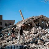 Zwei hagere Männer steigen über einen Trümmerberg, auf denen man noch Reste von Betonpfeilern und dem Dach eines Hauses erkennt