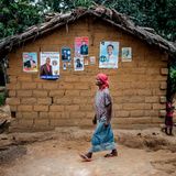 Eine schwarze Frau mit langem blauen Rock und rotem Kopftuch geht an einer Lehm-Hütte vorbei, an der Wahlplakate hängen
