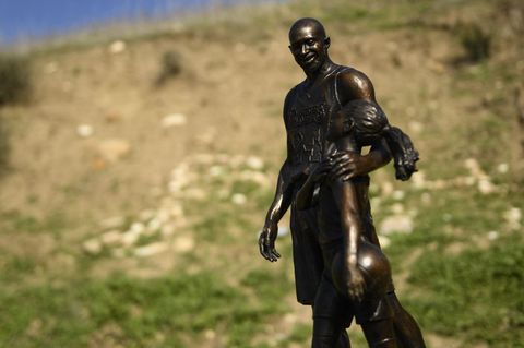 Vor einer teilweise mit Gras bewachsenen Abbruchkante steht die Statue eines Basketballers, der seinen Arm um ein Mächen legt