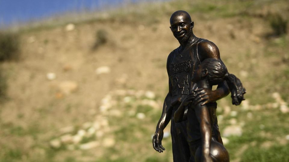Vor einer teilweise mit Gras bewachsenen Abbruchkante steht die Statue eines Basketballers, der seinen Arm um ein Mächen legt