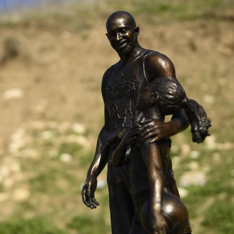 Vor einer teilweise mit Gras bewachsenen Abbruchkante steht die Statue eines Basketballers, der seinen Arm um ein Mächen legt