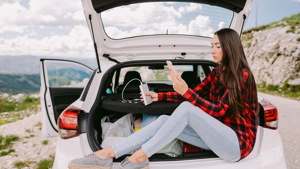 Eine junge Frau sitzt im Kofferraum eines Autos und lädt ihr Smartphone mit einer Powerbank