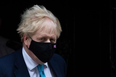 Boris Johnson kommt in Anzug und Krawatte aus einem dunklen Hausflur ins Freie