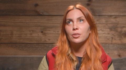 Eine junge weiße Frau mit langem orange-roten Haar sitzt vor einer Bretterwand und verdreht ihre blauen Augen