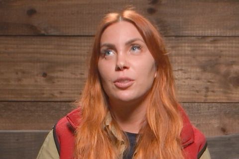 Eine junge weiße Frau mit langem orange-roten Haar sitzt vor einer Bretterwand und verdreht ihre blauen Augen