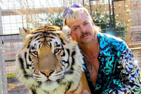 Ein weißer Mann mit blondiertem Vokuhila und Schnauzbart schmiegt sich an einen Tiger