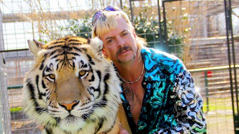 Ein weißer Mann mit blondiertem Vokuhila und Schnauzbart schmiegt sich an einen Tiger