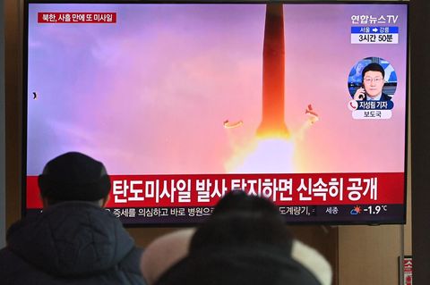 Während auf einem Fernseher eine startende Rakete und Einblendungen in koreanischer Schrift zu sehen sind, schauen Passanten zu