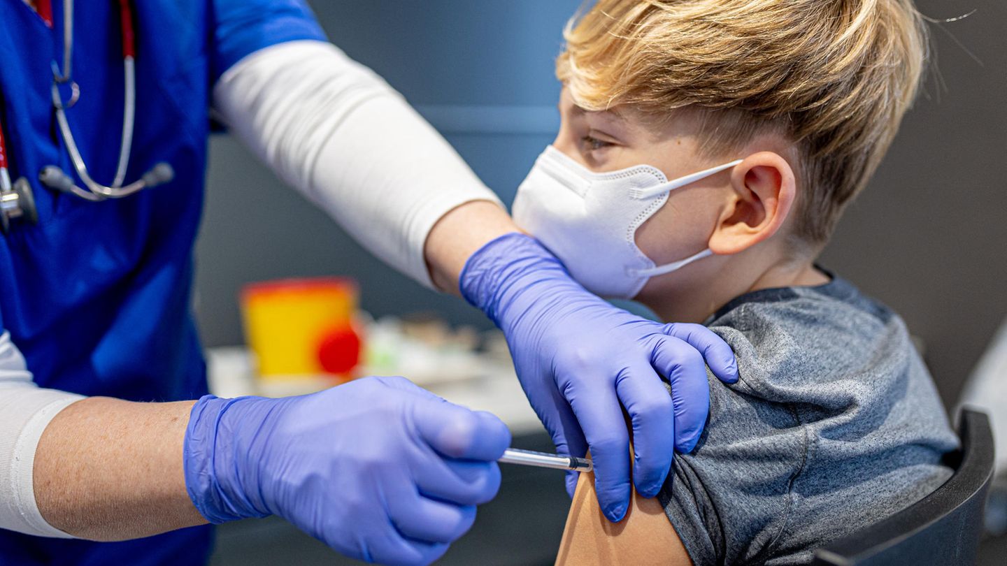 Ein weißer Junge mit blondem Seitenscheitel bekommt von einer Person in blauer medizinischer Kleidung eine Spritze verabreicht