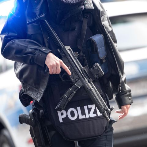 Vor einem blau-silbernen Polizeiauto steht ein Polizist in Schutzweste und mit Maschinengewehr vor dem Oberkörper