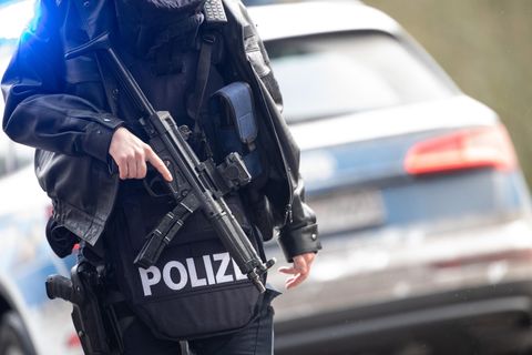Vor einem blau-silbernen Polizeiauto steht ein Polizist in Schutzweste und mit Maschinengewehr vor dem Oberkörper